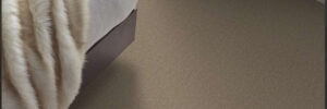 A bed with a white blanket and fur throw is positioned on light brown carpet next to a white nightstand.